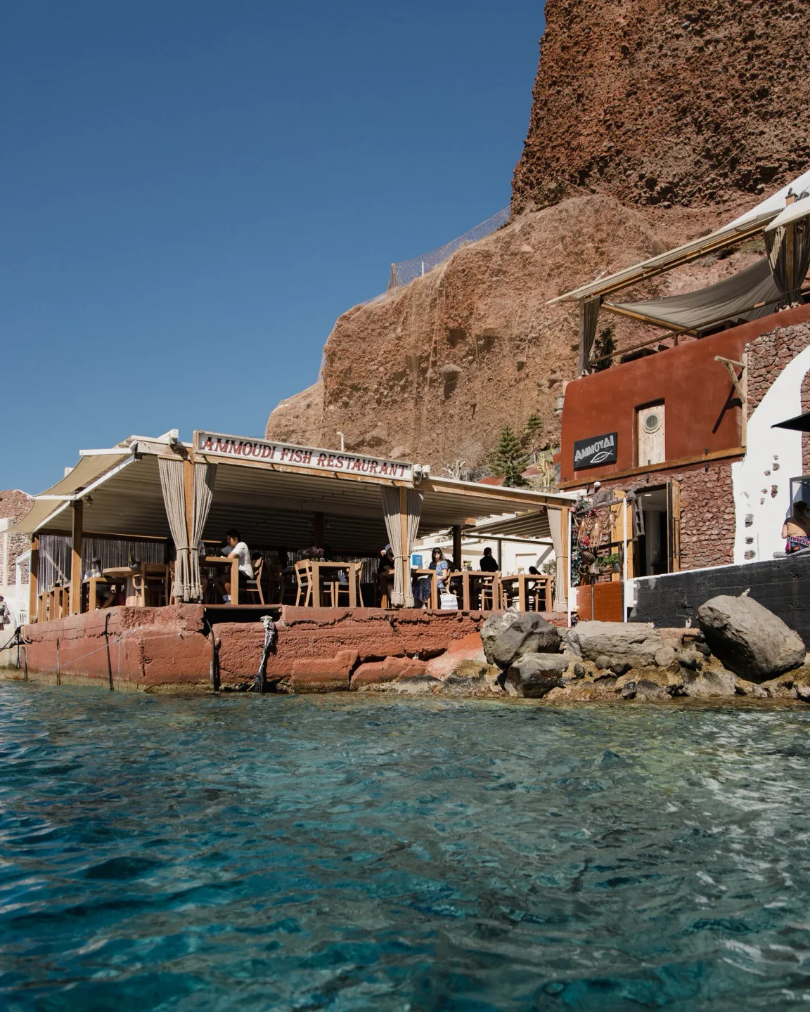 Ammoudi Fish Tavern, a waterfront restaurant in the city, with tables by the turquoise sea and rocky cliffs.