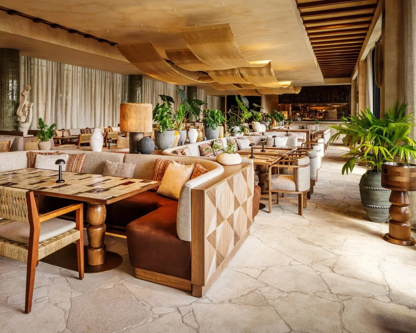 Casa Neos restaurant in Miami features warm lighting, lush plants, and comfortable booth seating with unique table designs.