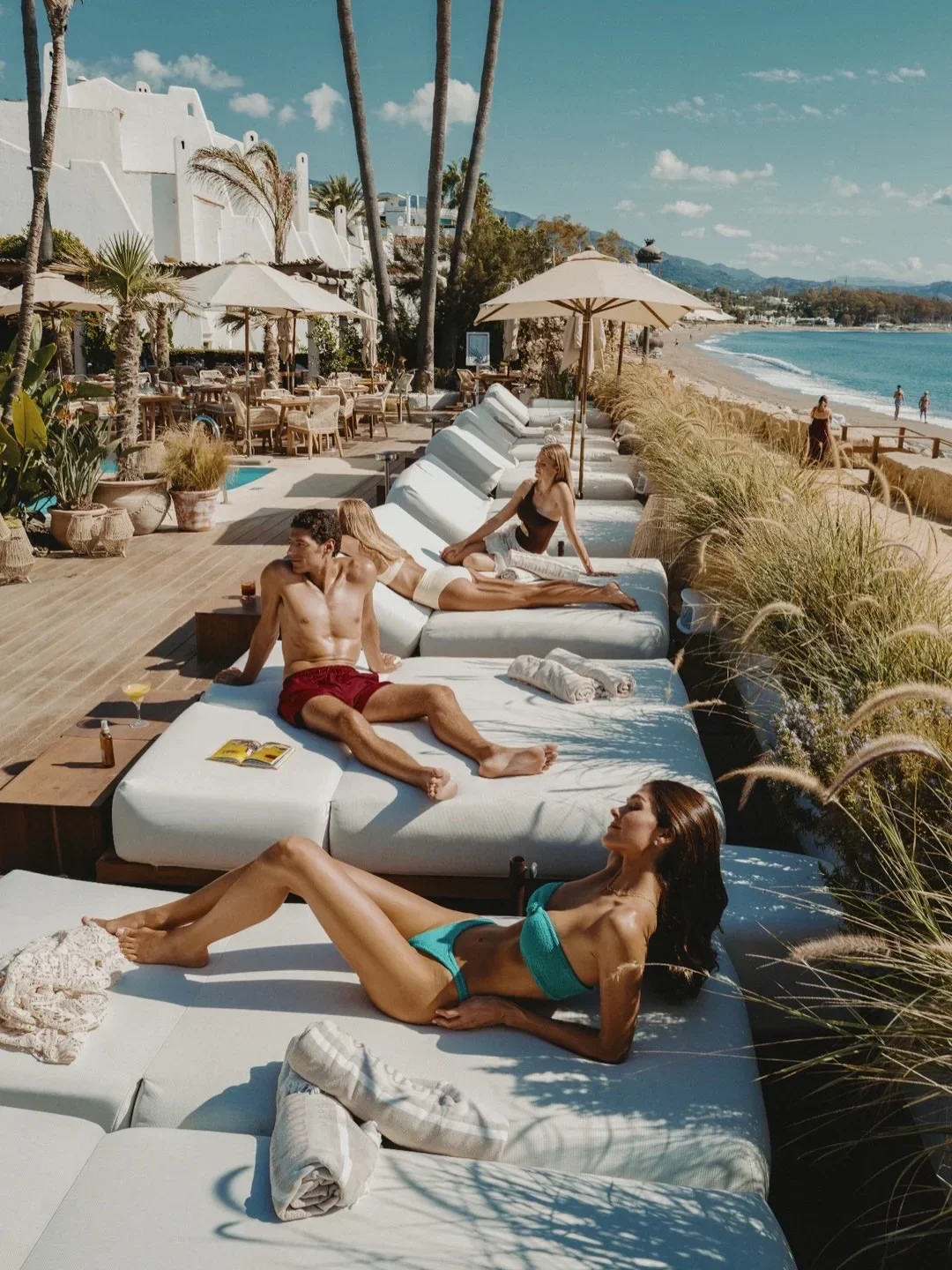 Couples relaxing on sun loungers at Chambao beach club in Marbella overlooking the Mediterranean Sea.