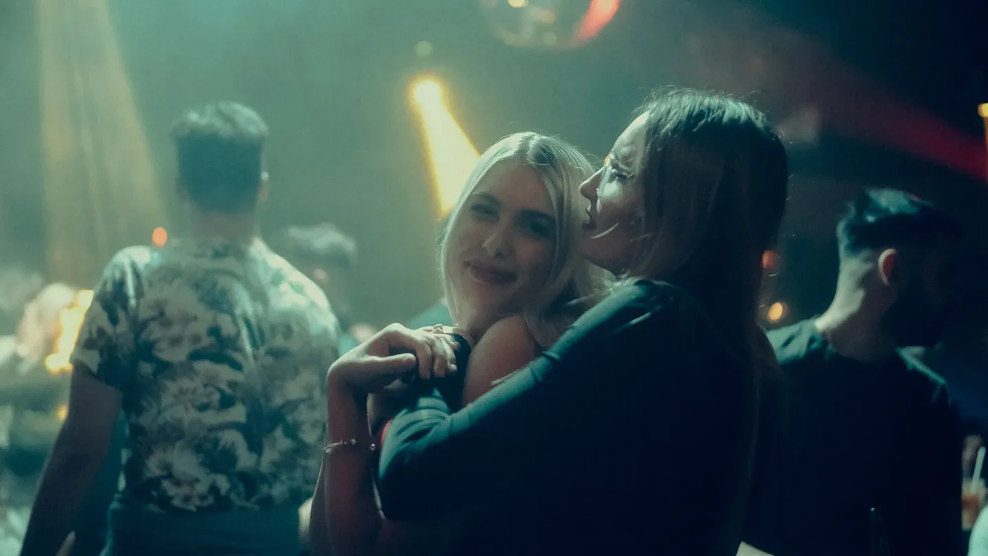 Two joyful women embracing on a lively dance floor at CLUB Badhanna Budapest, with colorful lights casting a vibrant glow.