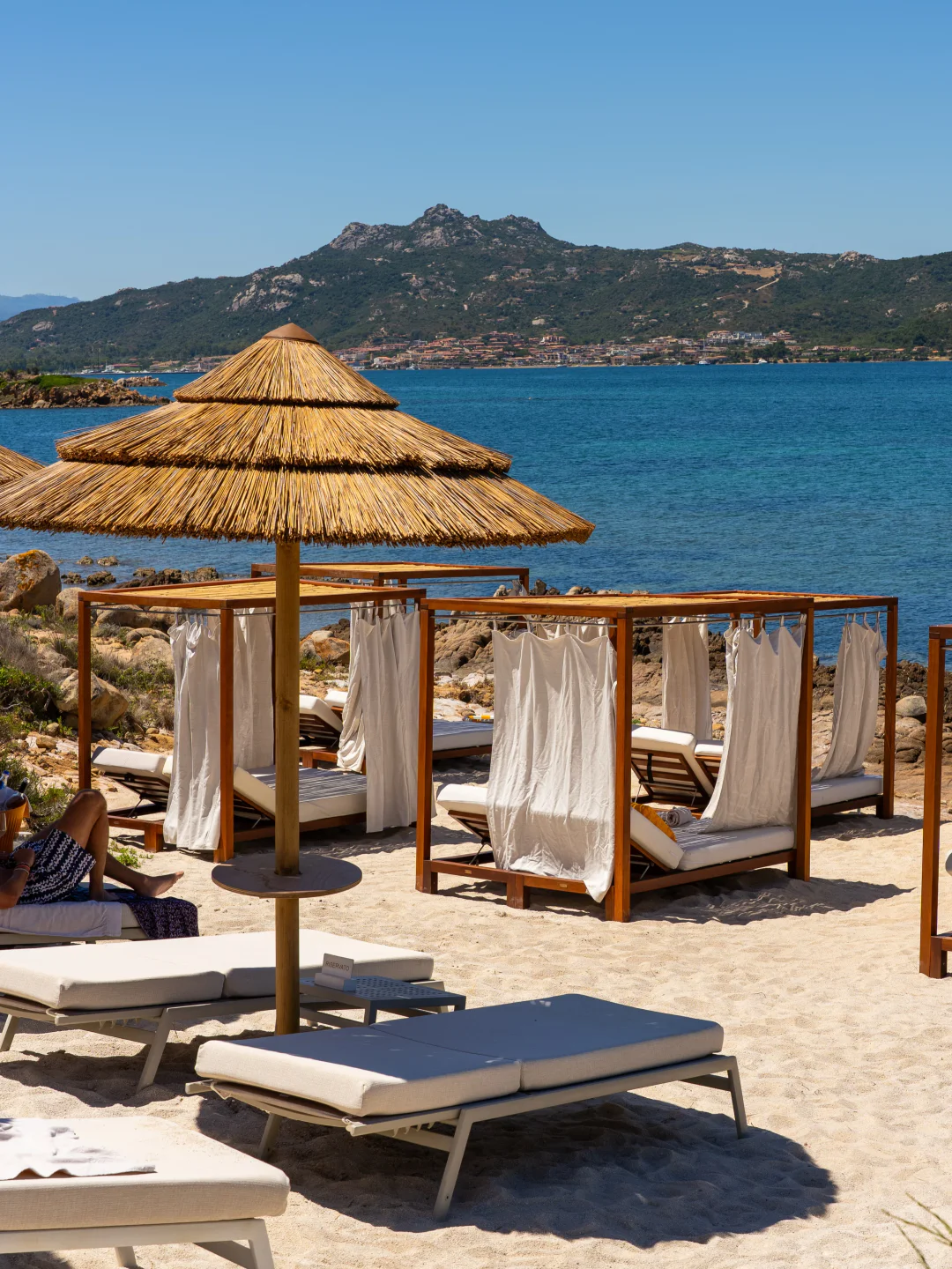 Cone Club Porto Cervo beach club with cabanas and lounge chairs by the turquoise sea and rocky coastline.