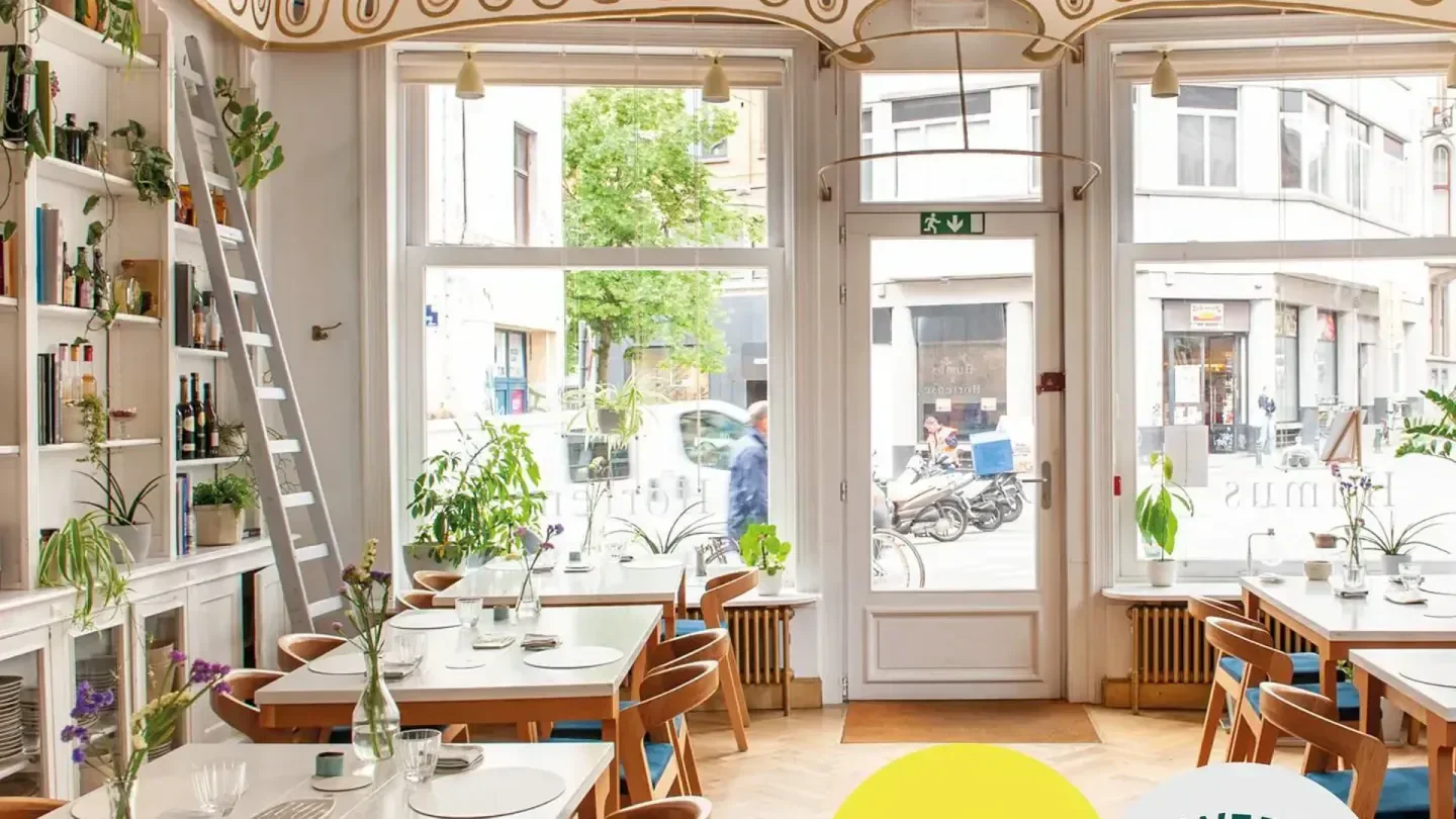 Interior of Humus x Hortense restaurant in Brussels, with tables, plants and street view.