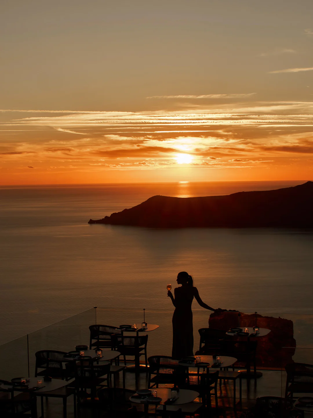 Silhouette of a woman holding wine at Mavro restaurant, overlooking the city and sea at sunset.