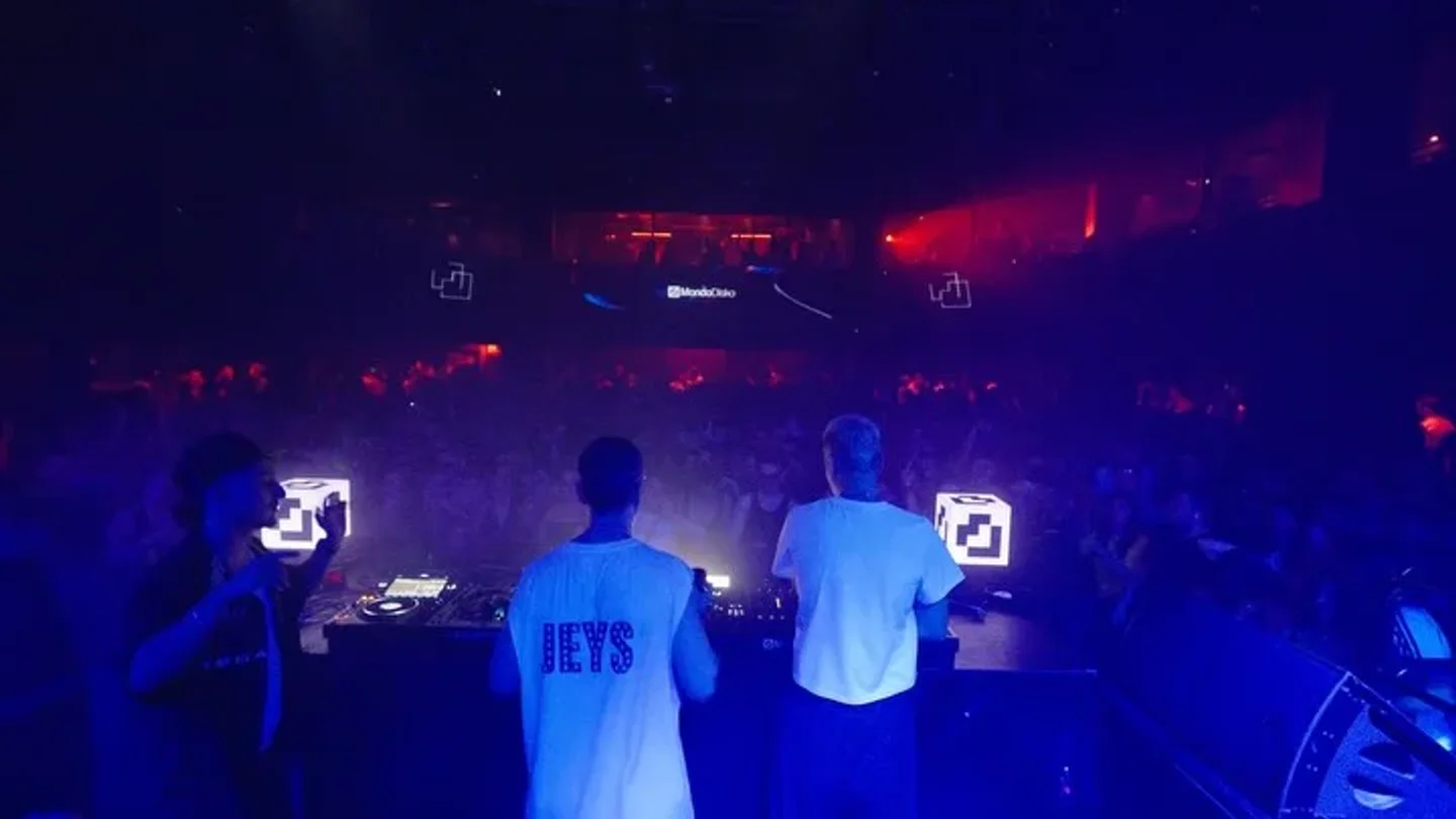 DJs perform for a crowd at Mondo Disko nightclub, illuminated by vibrant blue and red lights.