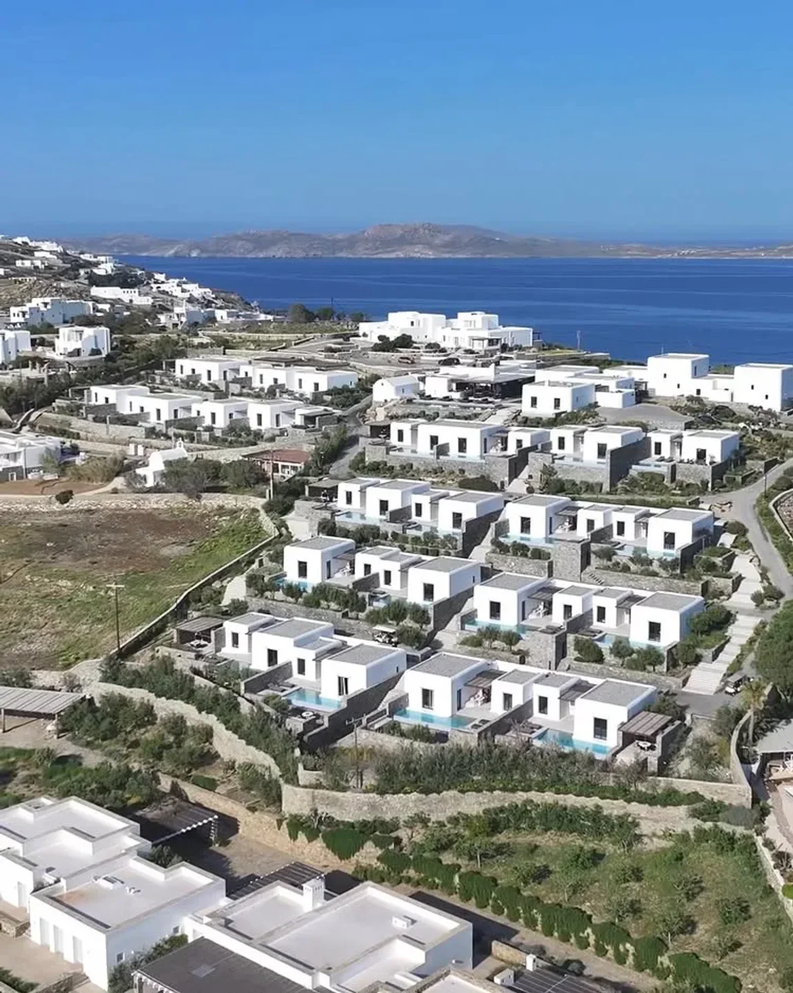 Hillside view of modern white villas with pools at Pere Ubu restaurant in Mykonos overlooking the Aegean Sea.