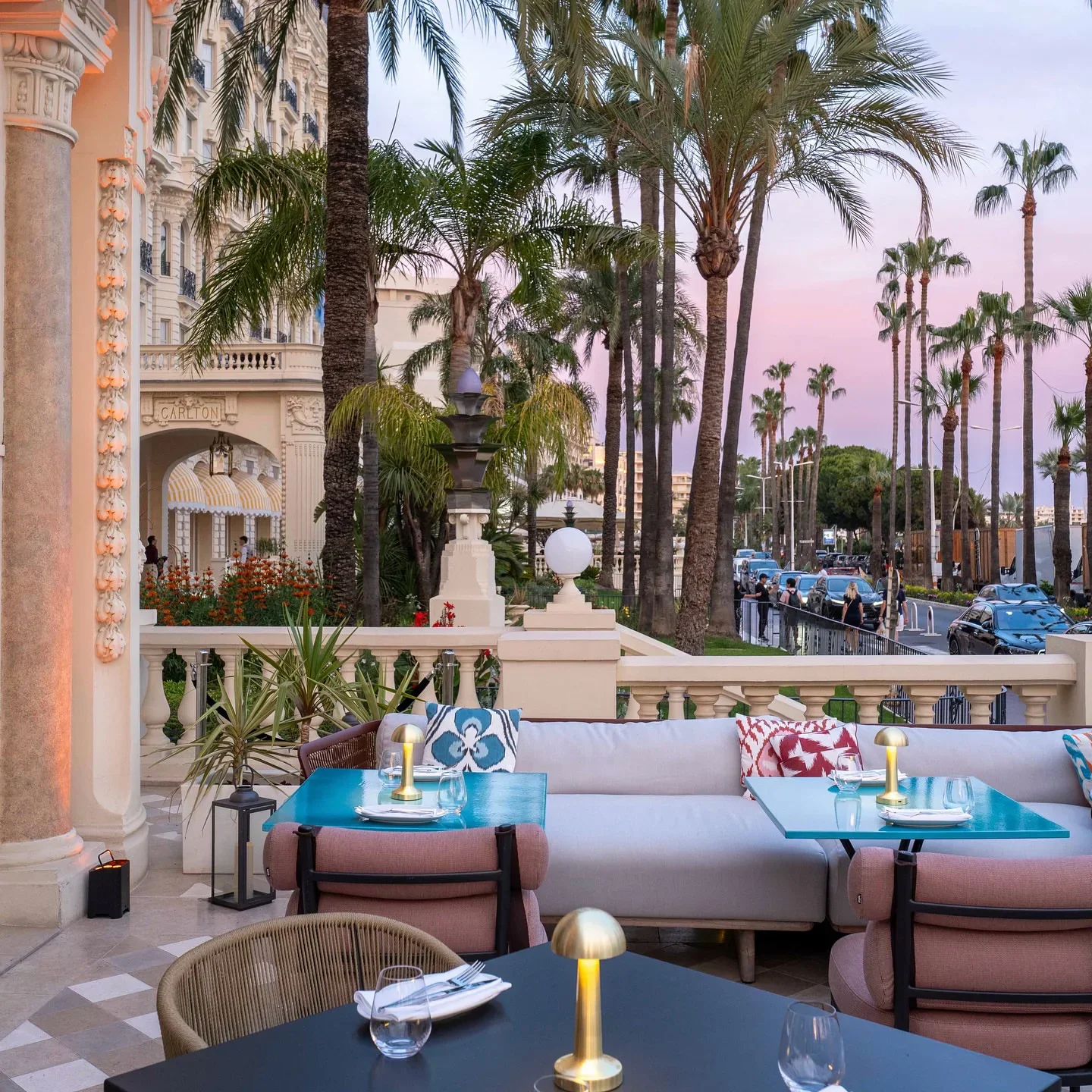 Outdoor seating area at Ruya restaurant in Cannes with palm trees and a pink sunset sky.