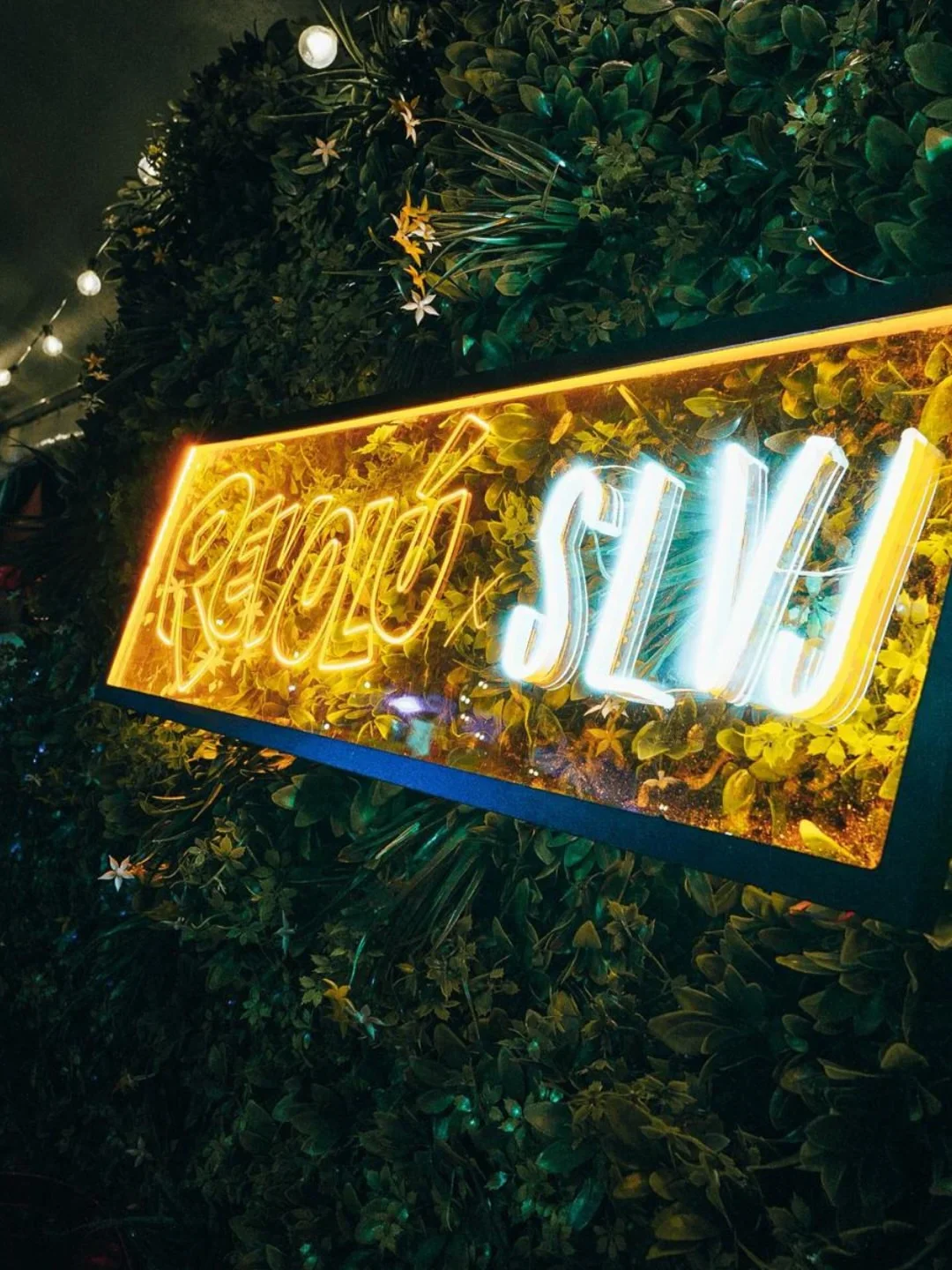 Neon "SLVJ" sign glowing against a lush green wall with fairy lights, hinting at the vibrant ambiance of the city restaurant.