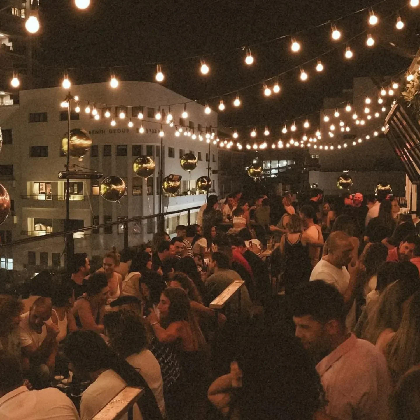 Speakeasy restaurant in Tel Aviv, crowded rooftop party under the string lights at night.