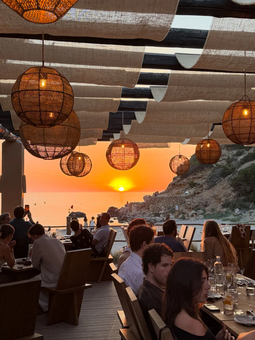 Romantic sunset view from Urusai restaurant overlooking the sea, with people dining under unique woven lamps.